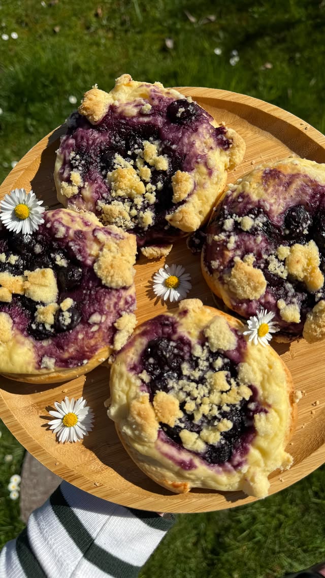 Vier Stücke hausgemachter Heidelbeer-Streuselkuchen auf einem Holzteller, dekoriert mit Gänseblümchen, im Gras fotografiert.