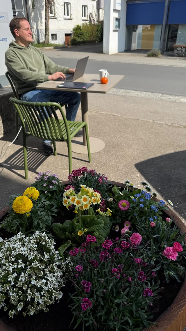 Draußen vor dem Café: jemand arbeitet am Laptop an einem kleinen Tisch auf einem grünen Stuhl, im Vordergrund ein großer Kübel voller bunter Frühlingsblumen.