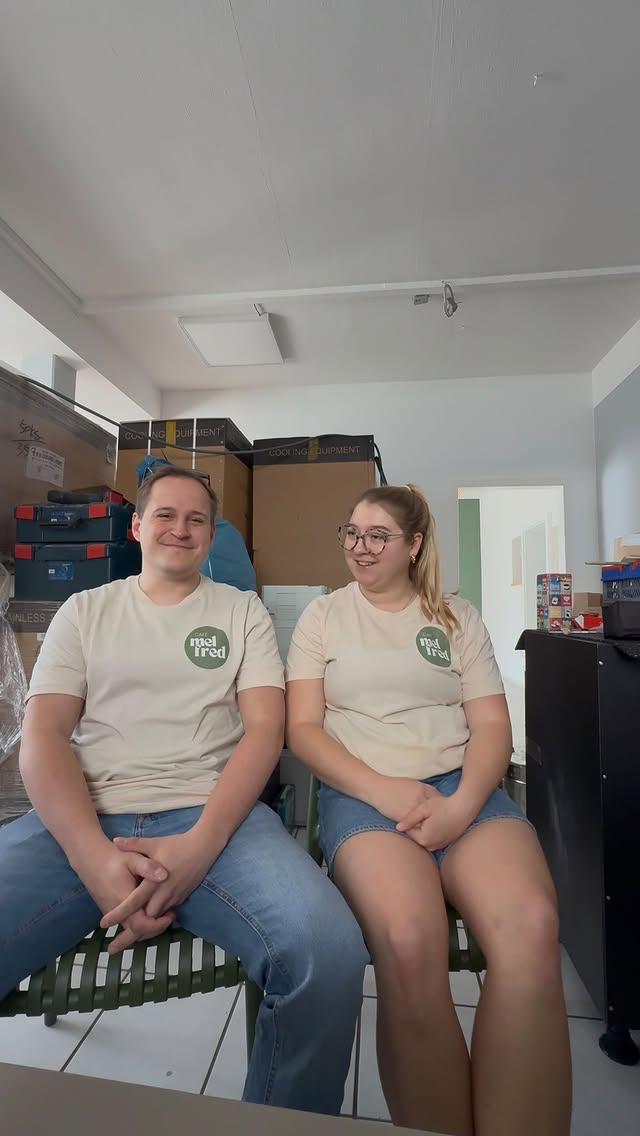 Die beiden Gründer:innen in „mel fred“-Shirts sitzen lächelnd zwischen Umzugskartons mit Café-Ausstattung.